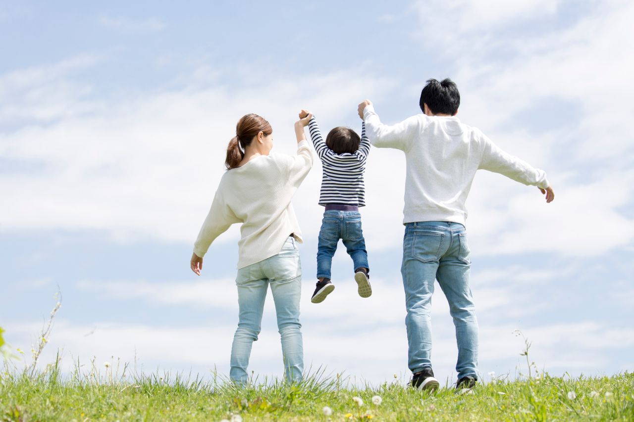 青空と草むらを背景に、若い夫婦が小さな子どもと手を繋いで楽しそうに歩いている家族の後ろ姿