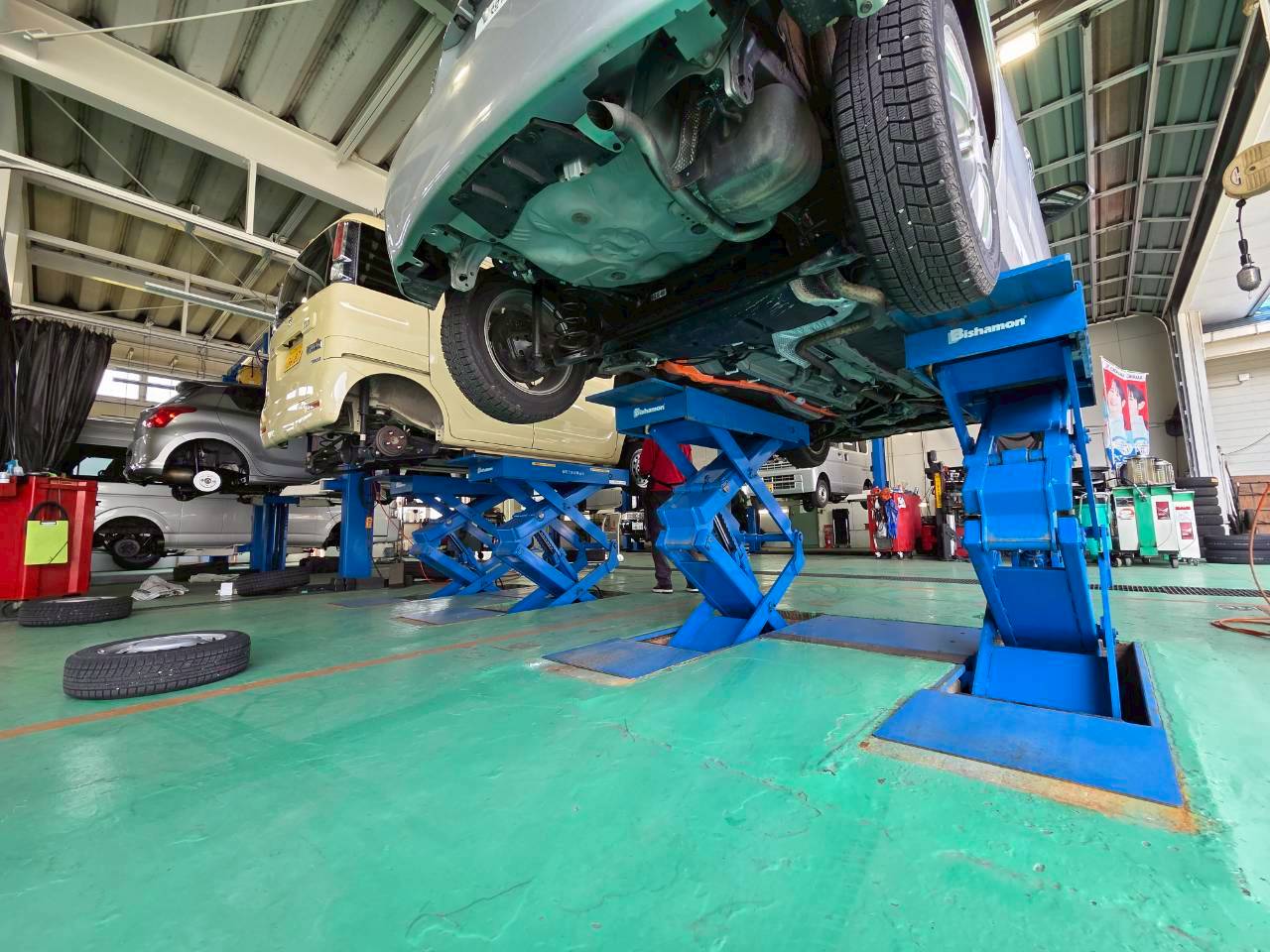 青いリフトで高く持ち上げられた車両を下から点検・整備している整備工場の様子