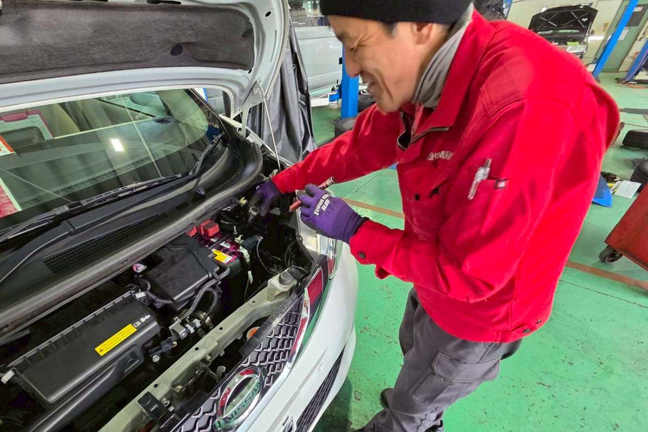 ボンネットを開けた車のエンジンルームを笑顔で点検している、赤いジャンパーを着た整備士