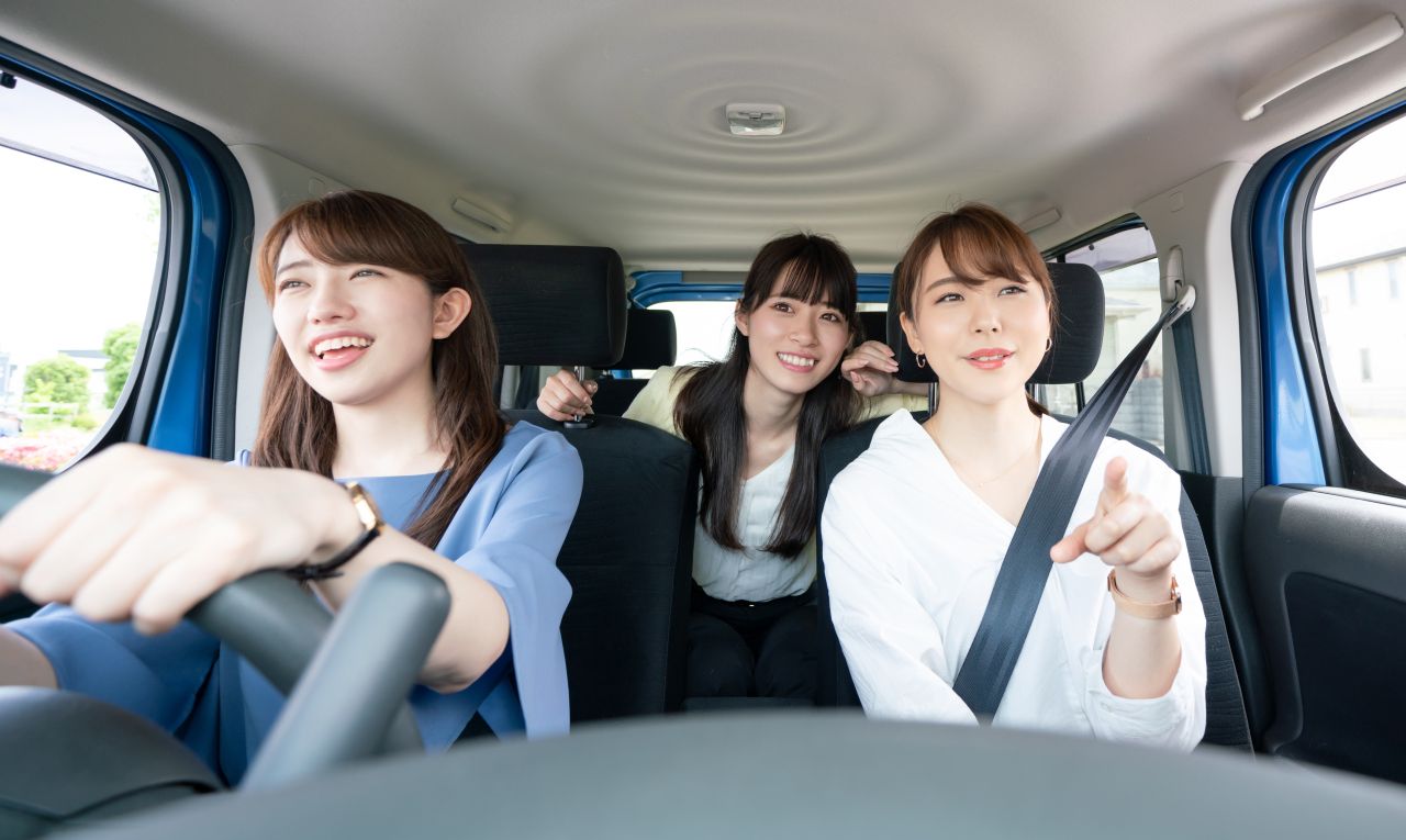 レンタカーの車内で、笑顔の女性3人が楽しそうにドライブをしている様子