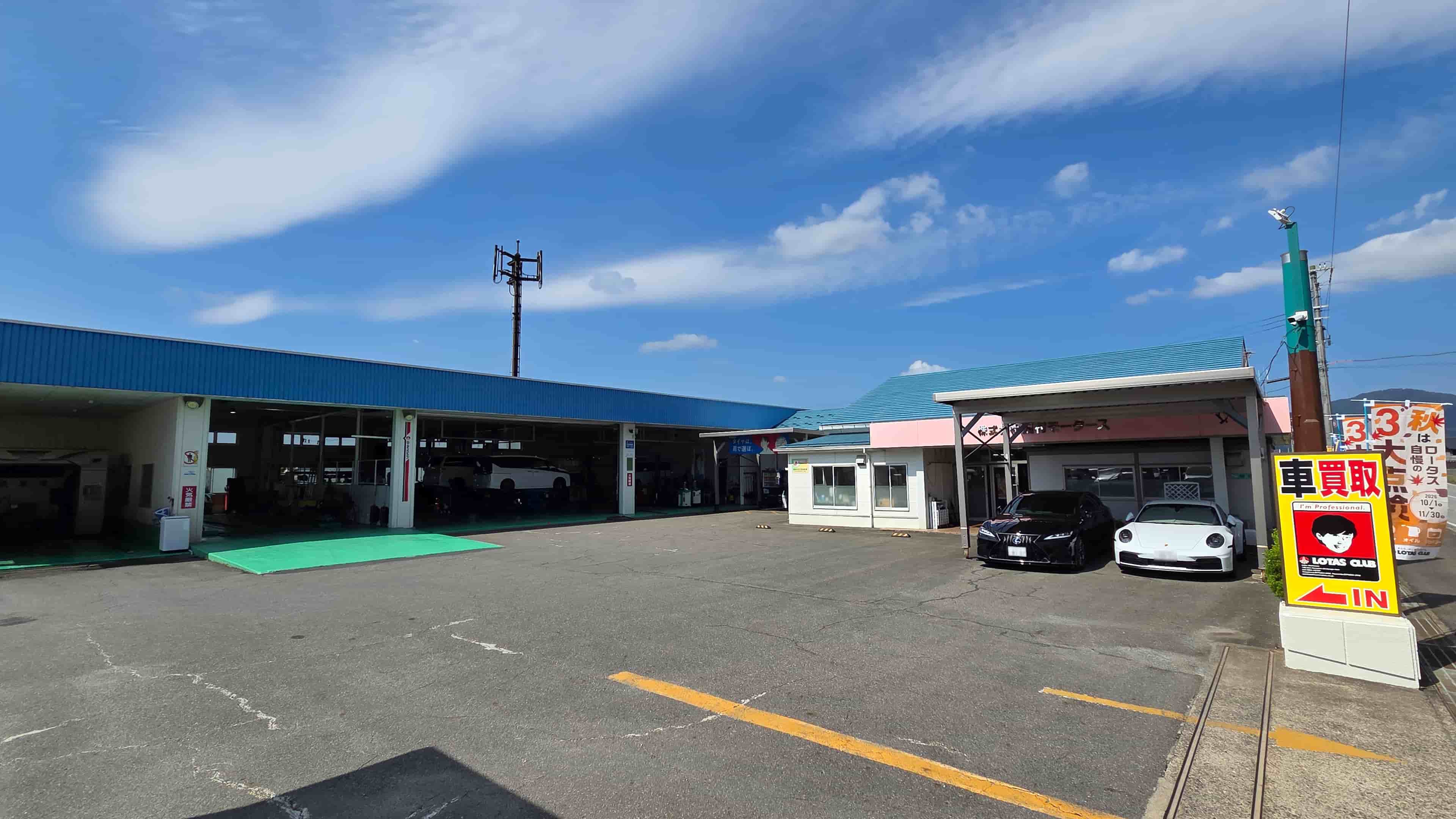 青空の下に広がる株式会社西村モータースの店舗外観。左側には洗車機や複数の作業ベイを備えた大きな整備工場、右側には受付オフィスと車両展示スペースがあり、手前には「車買取」の看板が立っている。