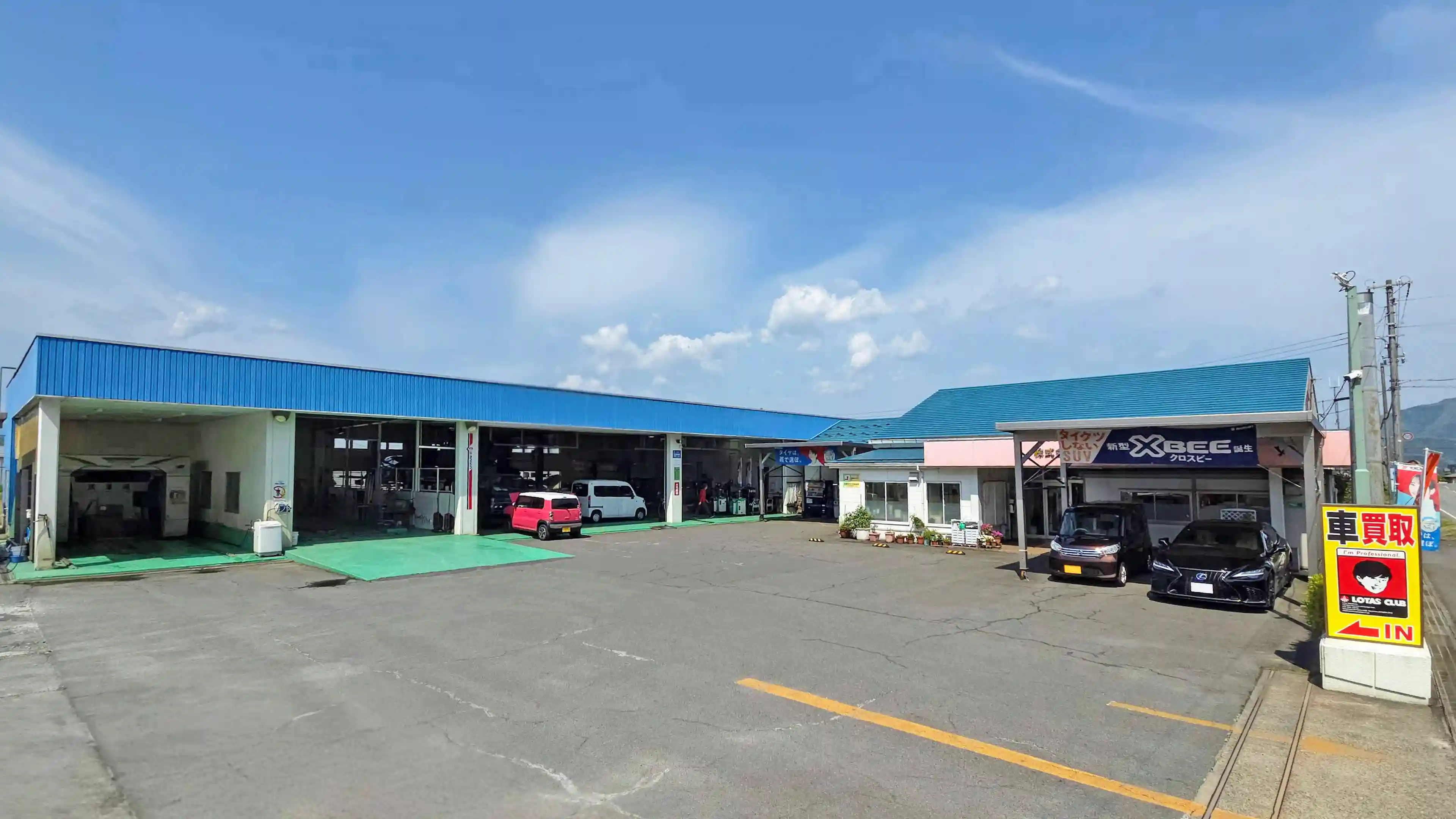 青空の下に広がる株式会社西村モータースの店舗外観。左側には洗車機や複数の作業ベイを備えた大きな整備工場、右側には受付オフィスと車両展示スペースがあり、手前には「車買取」の看板が立っている。