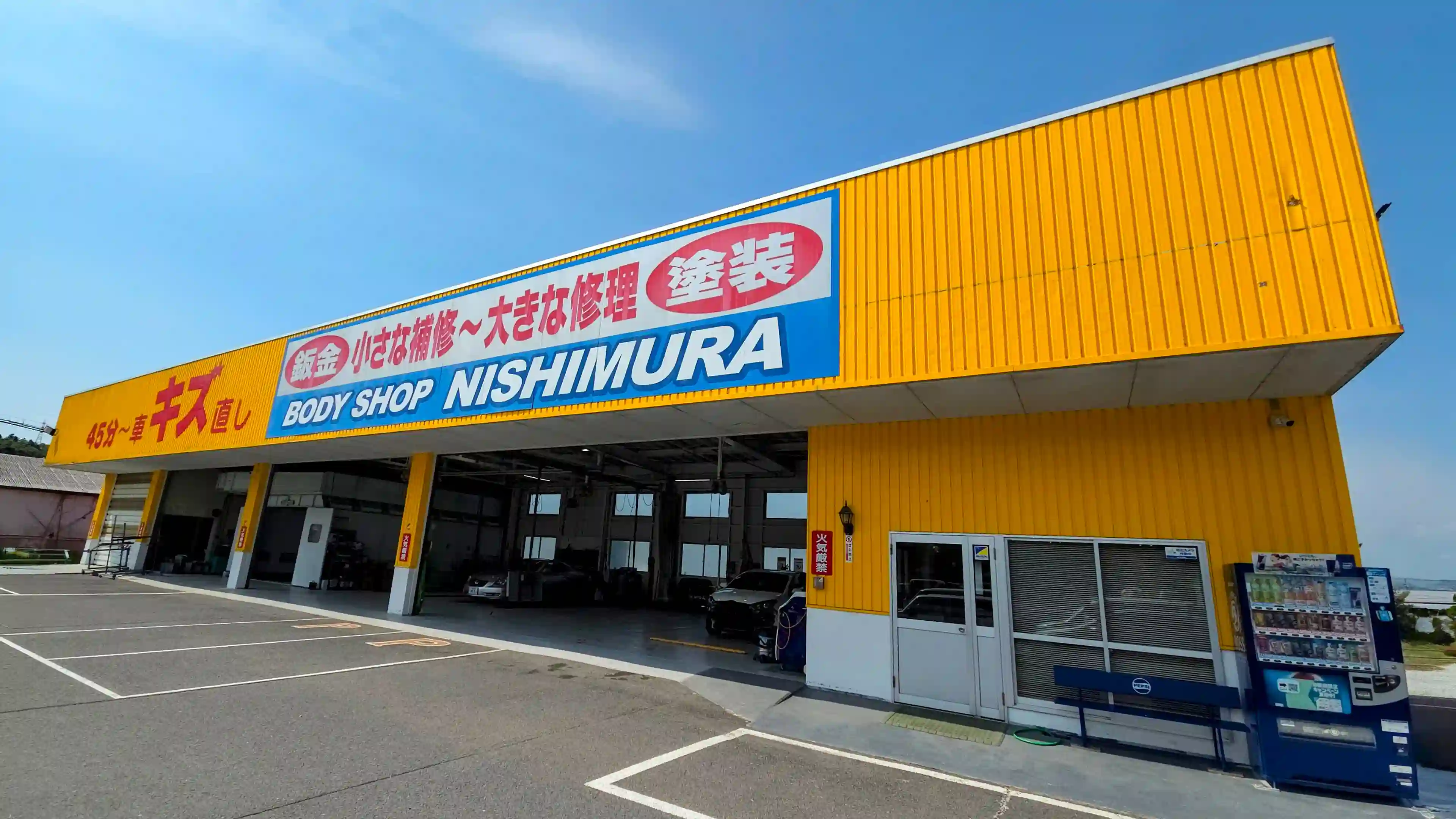 鮮やかな黄色の壁が特徴的な「BODY SHOP NISHIMURA」の工場外観。「45分〜車のキズ直し」「鈑金 小さな補修〜大きな修理 塗装」と大きく書かれた看板の下に、複数の作業ピットが並び、内部に車が格納されている様子
